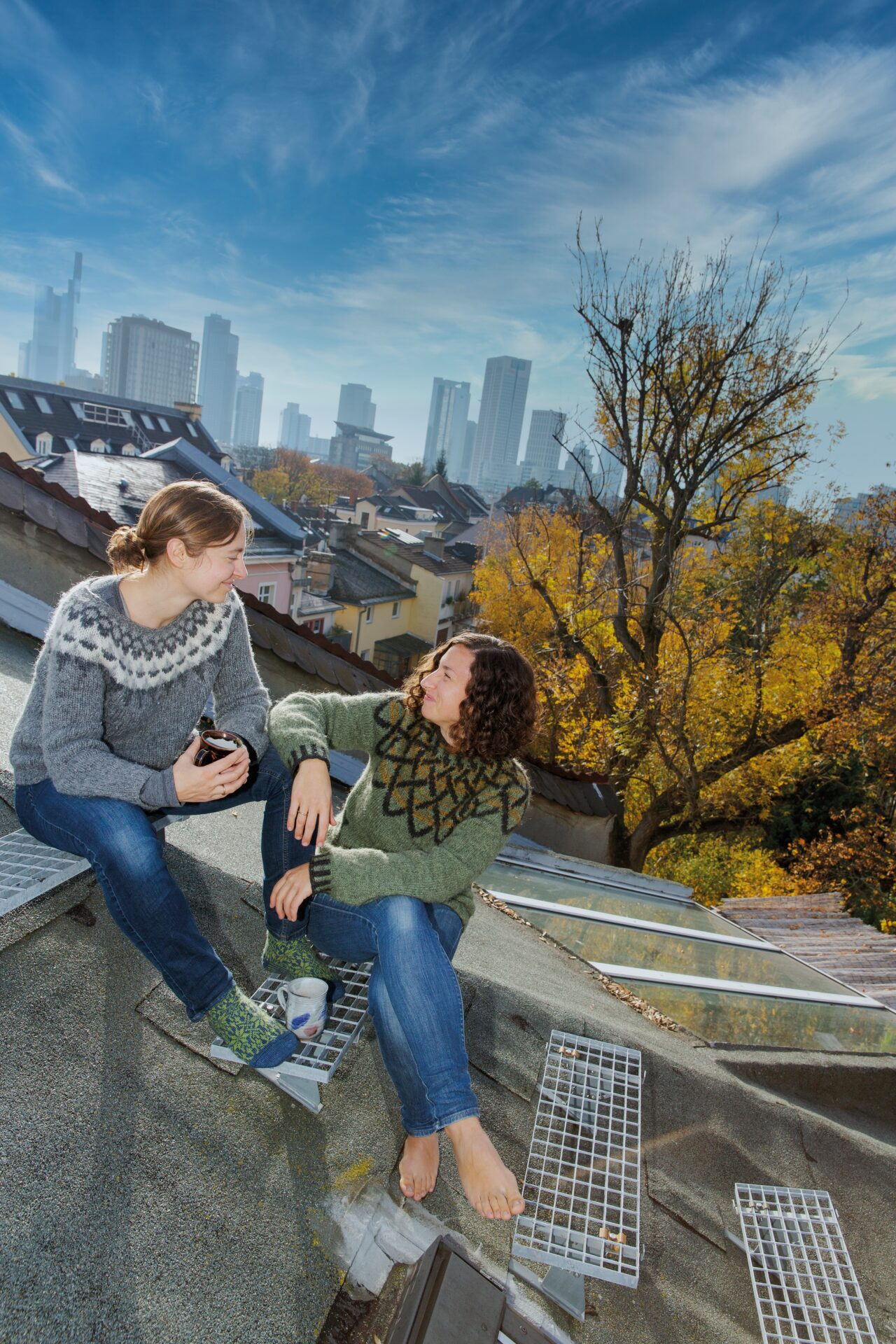 Die Autorinnen Juliane und Laura im Gespräch, auf einem Stadtdach sitzend, die Skyline im Hintergrund.