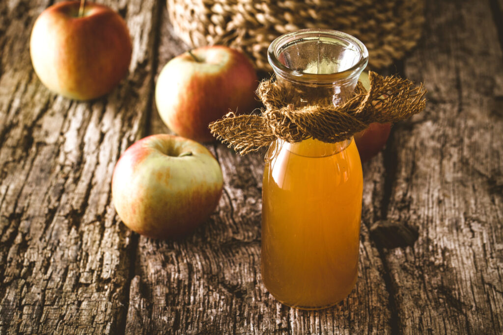 Apfelessig als Spülung: Naturtrüber Apfelessig steht in einem Glasgefäß auf einem Holztisch neben Äpfeln