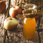 Apfelessig als Spülung: Naturtrüber Apfelessig steht in einem Glasgefäß auf einem Holztisch neben Äpfeln