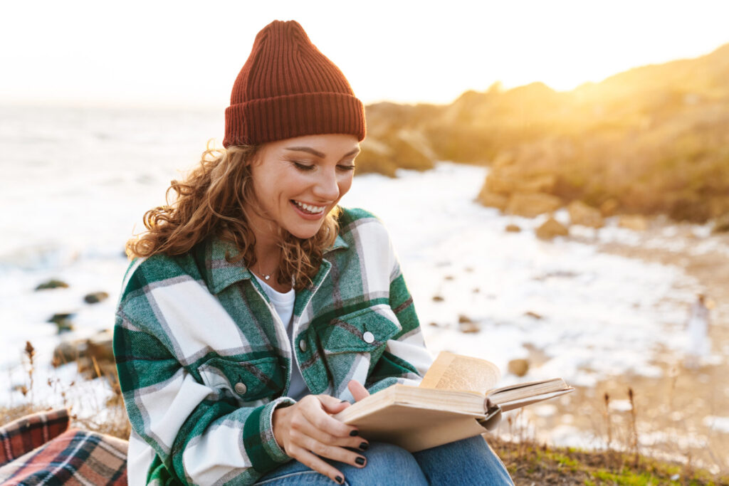 Heilfasten nach Buchinger macht glücklich: eine fröhliche junge Frau liest ein Buch an der Küste.